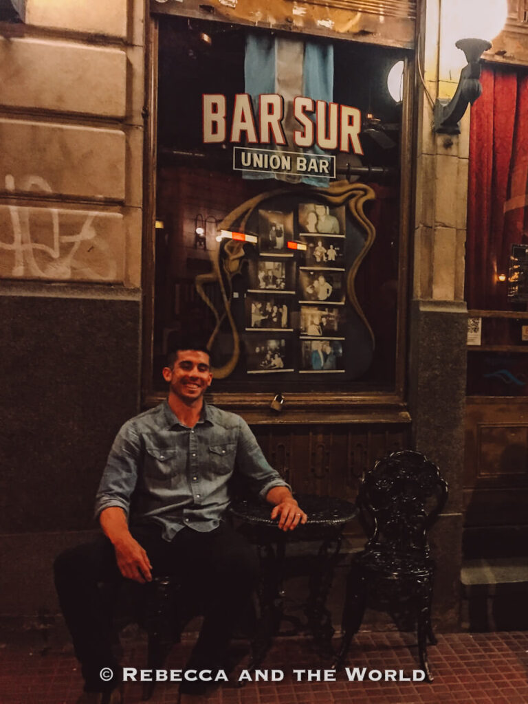A man sits on a chair outside a venue at night, smiling toward the camera. Behind him, a window display with framed photos and a sign reading “Bar Sur Union Bar” is visible. Make sure you go to a tango show in Buenos Aires, Argentina.