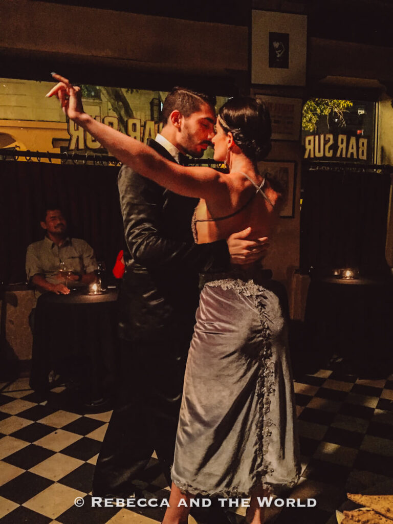 A couple performs a tango dance in a dimly lit room. The man wears a dark suit while the woman wears a fitted dress, and they hold each other closely as they dance. Other patrons sit at tables in the background. This is Bar Sur in Buenos Aires, Argentina.