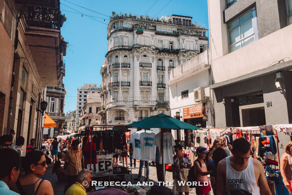 A busy outdoor market scene with people walking between stalls selling clothing and goods. Historic buildings line the street, and the sky is bright and clear. The Weekly Feria de San Telmo is one of the best markets in Buenos Aires.
