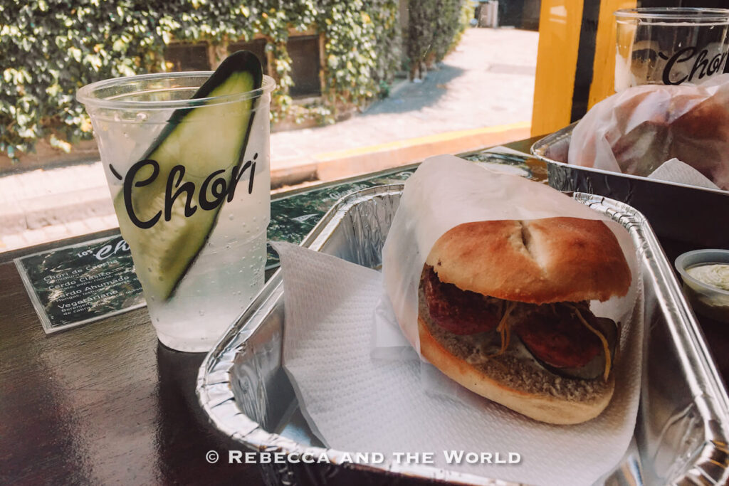 A casual meal on a table includes a choripan served in a foil tray and a clear plastic cup with a drink and a slice of cucumber inside. Another tray with food is partially visible nearby. Choripan is one of the best Argentina foods to try.