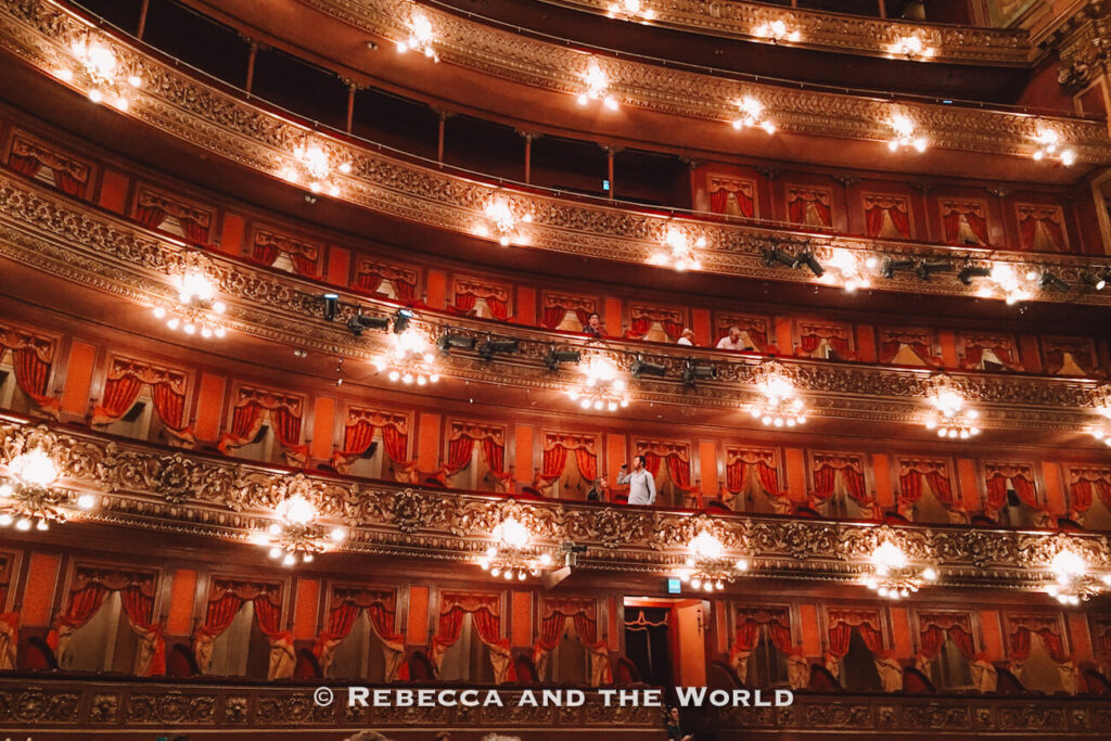 The interior of a grand theater with multiple balconies lined with red curtains and ornate gold detailing. Several people stand along the railings, looking out over the seating area below. Try to get tickets for a show at the Teatro Colon on your 3 days in Buenos Aires - or just add a guided tour to your itinerary.