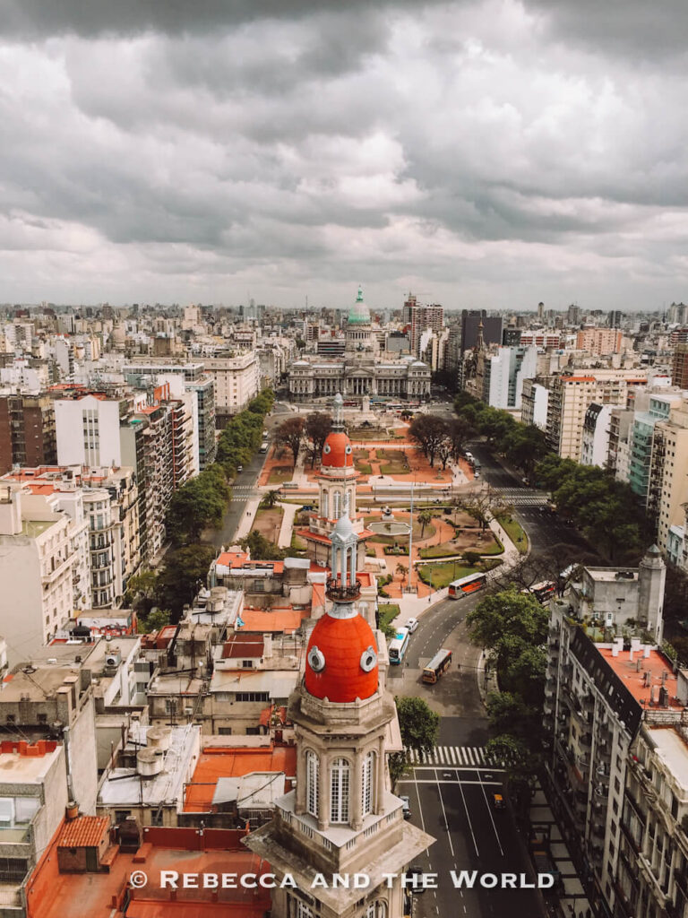 An elevated view of a large city avenue lined with mid-rise buildings. In the foreground, a historic tower with a red dome stands above the street, while farther back a grand government building with a green dome faces a plaza. The sky is overcast with thick gray clouds. This is the view of Buenos Aires from the top of Palacio Barolo.