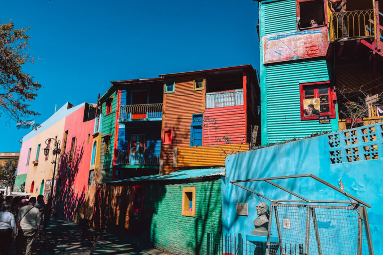 Colorful houses in La Boca with bright blue, red, yellow, and green facades, balconies, and open windows. Several people walk along the street below, looking up at the buildings. This lively street scene reflects popular things to do in Buenos Aires and is often included in a 3 days in Buenos Aires itinerary.