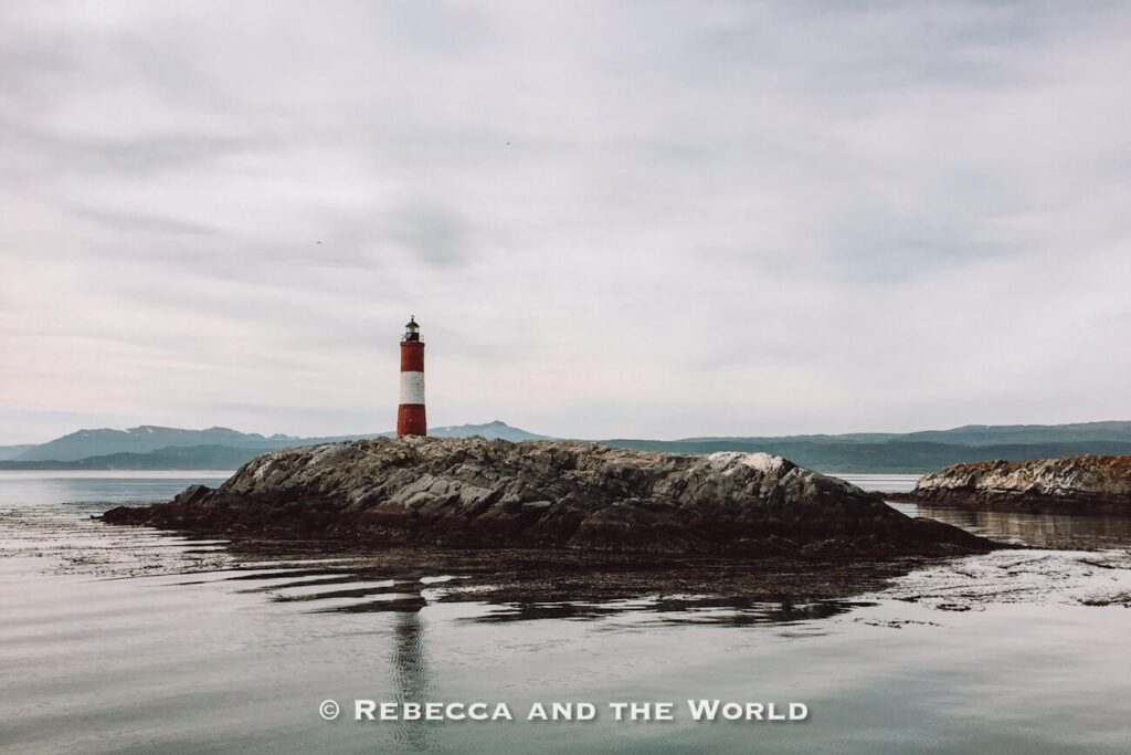 Red and white striped lighthouse standing on a rocky islet surrounded by calm water under an overcast sky. Low mountains stretch across the horizon in the background. Going to Ushuaia - the "end of the world" is one of the best things to do in Argentina.