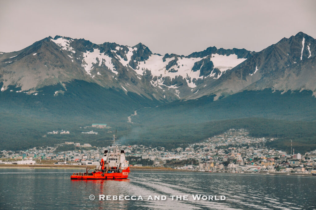 Red and white striped lighthouse standing on a rocky islet surrounded by calm water under an overcast sky. Low mountains stretch across the horizon in the background. Going to Ushuaia - the "end of the world" is one of the best things to do in Argentina.