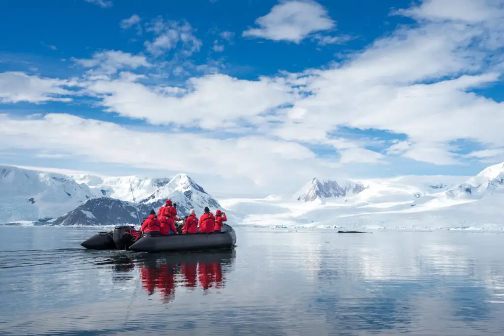 The image shows a group of people dressed in vibrant red expedition jackets, seated in an inflatable boat as they glide across a calm, reflective body of water. They are surrounded by a breathtaking icy landscape with snow-covered mountains and glaciers under a bright blue sky scattered with fluffy white clouds. The scene conveys the pristine beauty and serene atmosphere of an Antarctic expedition.