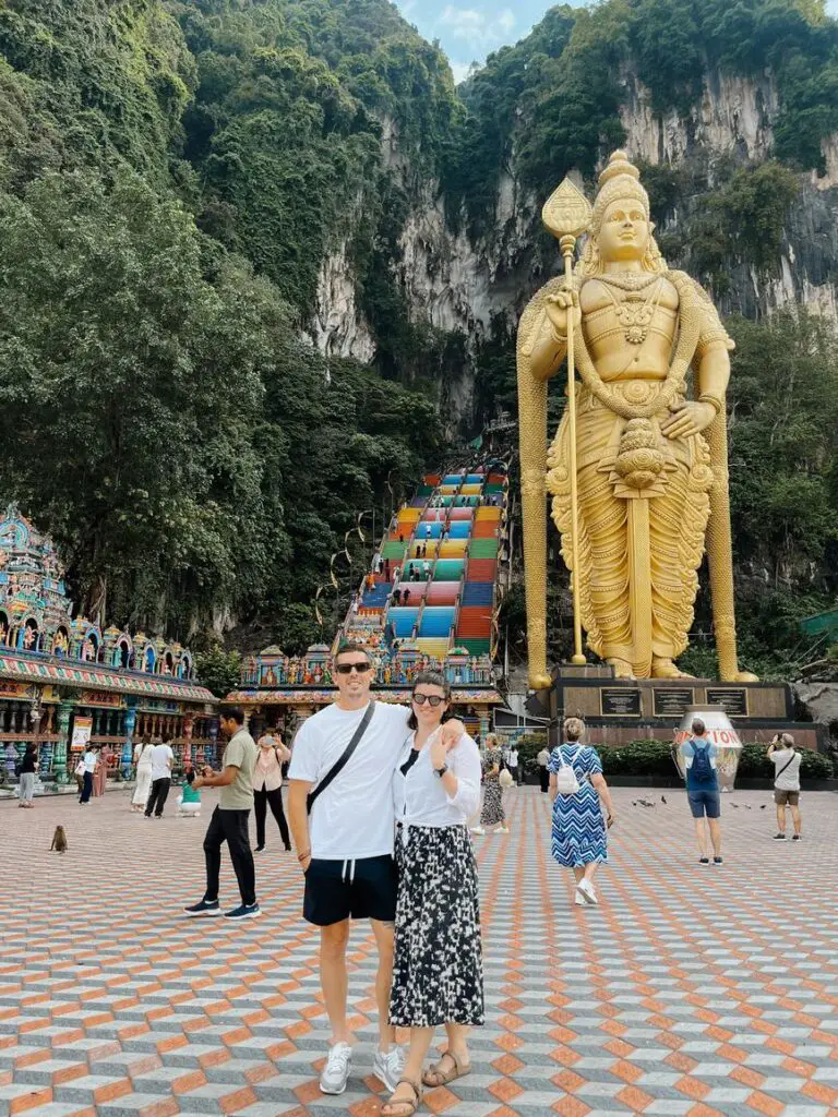 A couple - the author and her husband - posing in front of the iconic golden statue of Lord Murugan and the vibrant rainbow staircase at Batu Caves, with steep limestone cliffs and temple structures behind them.
