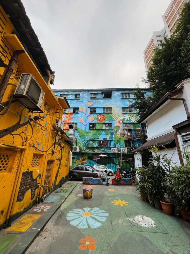 A vibrant alleyway in Bukit Bintang, Kuala Lumpur, with brightly painted yellow and blue buildings, colorful murals, and floral street art on the pavement.