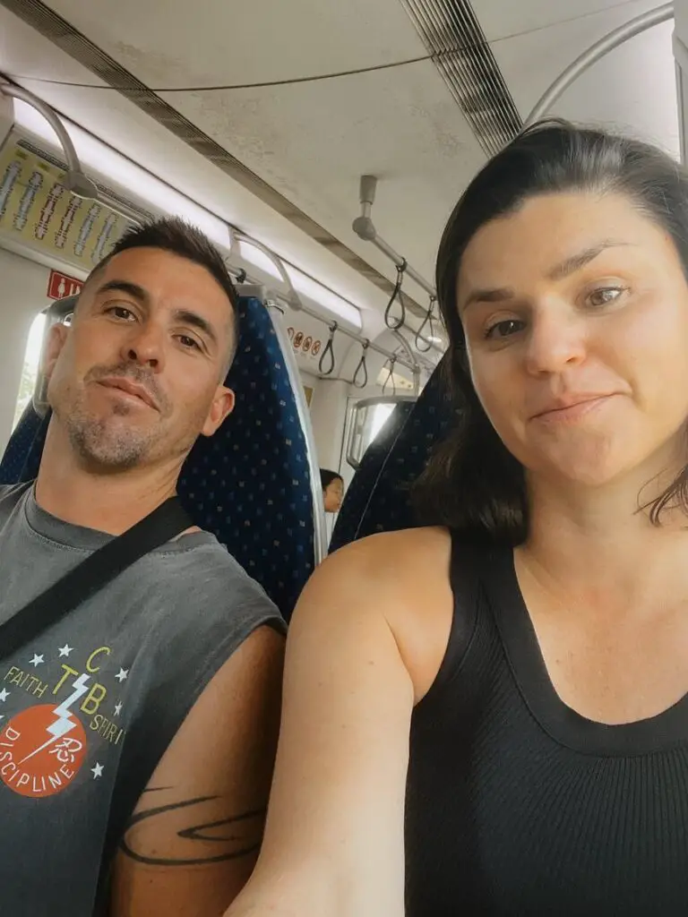 Selfie of a couple - the author and her husband - seated inside a commuter train in Malaysia, with overhead handles and passengers in the background.