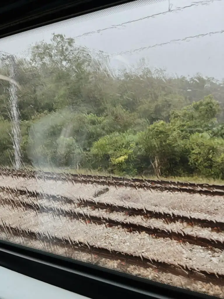 View through a rain-streaked train window looking out at railway tracks and a lush green forest during a journey from Gemas to Kuala Lumpur.