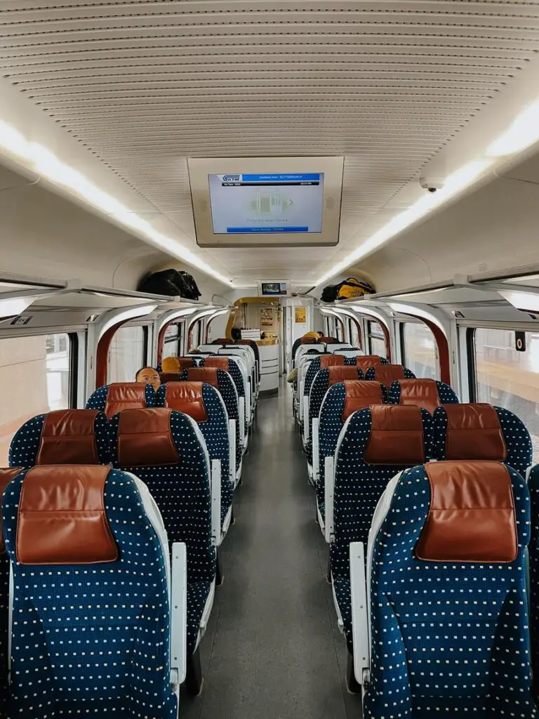 Inside a modern KTM ETS train in Malaysia, showing rows of blue and white polka-dotted seats with leather headrests, luggage racks above, and a digital screen displaying route information.