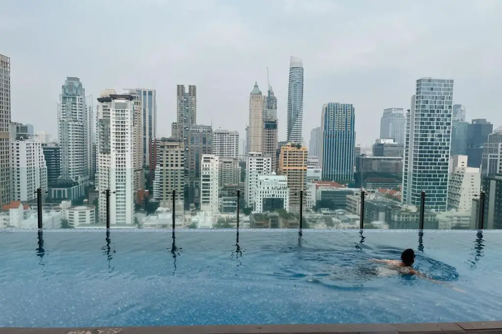 View of a man - the author's husband - swimming in a rooftop infinity pool with the Bangkok skyline rising in the background on a hazy day.