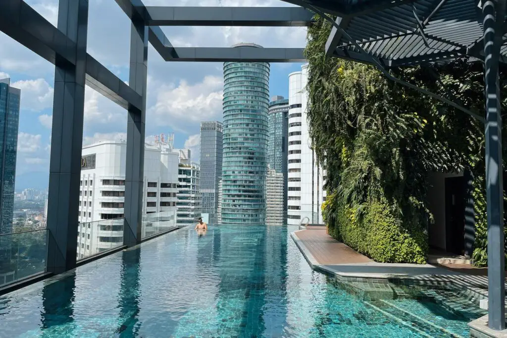 Infinity rooftop pool at Hotel Indigo Kuala Lumpur with a lush vertical garden on one side and a view of modern city skyscrapers and blue sky in the background. The author's husband is in the pool.