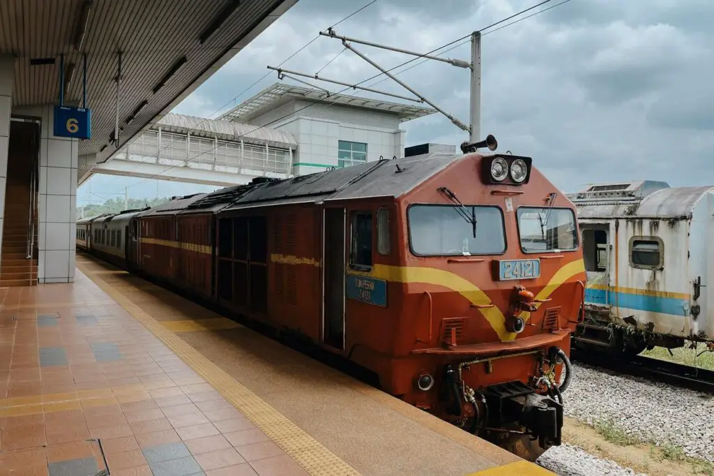A red and yellow diesel locomotive at Gemas station in Malaysia, parked next to a platform with a covered pedestrian bridge in the background under cloudy skies.