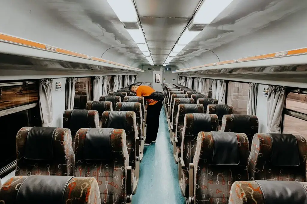 Interior of a Malaysian long-distance train with rows of cushioned seats arranged in a 2x2 layout, curtains along the windows, and a passenger in an orange shirt stowing luggage.