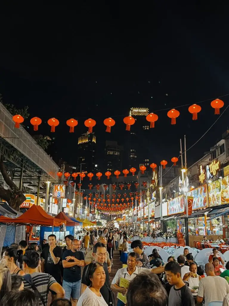 Bustling night market at Jalan Alor in Kuala Lumpur, packed with people and illuminated by rows of red Chinese lanterns hanging above the crowded street.