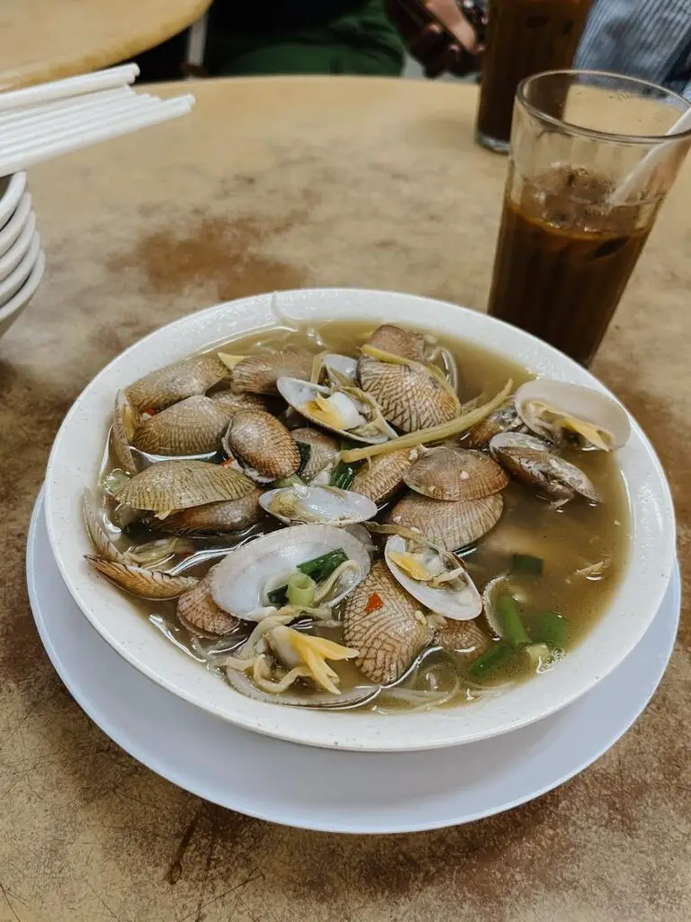 Bowl of hot and spicy clam soup with shell-on clams, ginger, chili, and green onions, served in a white ceramic bowl at a street food stall in Kuala Lumpur.