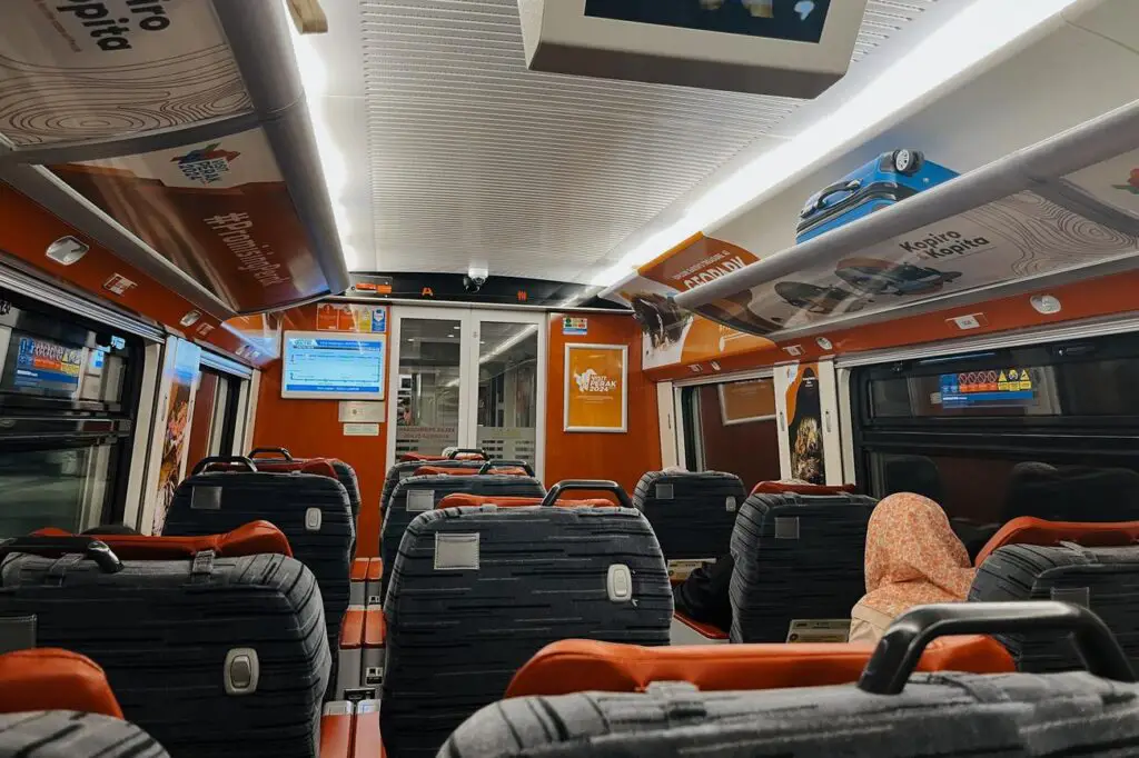Interior of a KTM ETS business class train carriage, showing rows of grey and orange seats, digital screens, and overhead luggage racks with promotional signage.