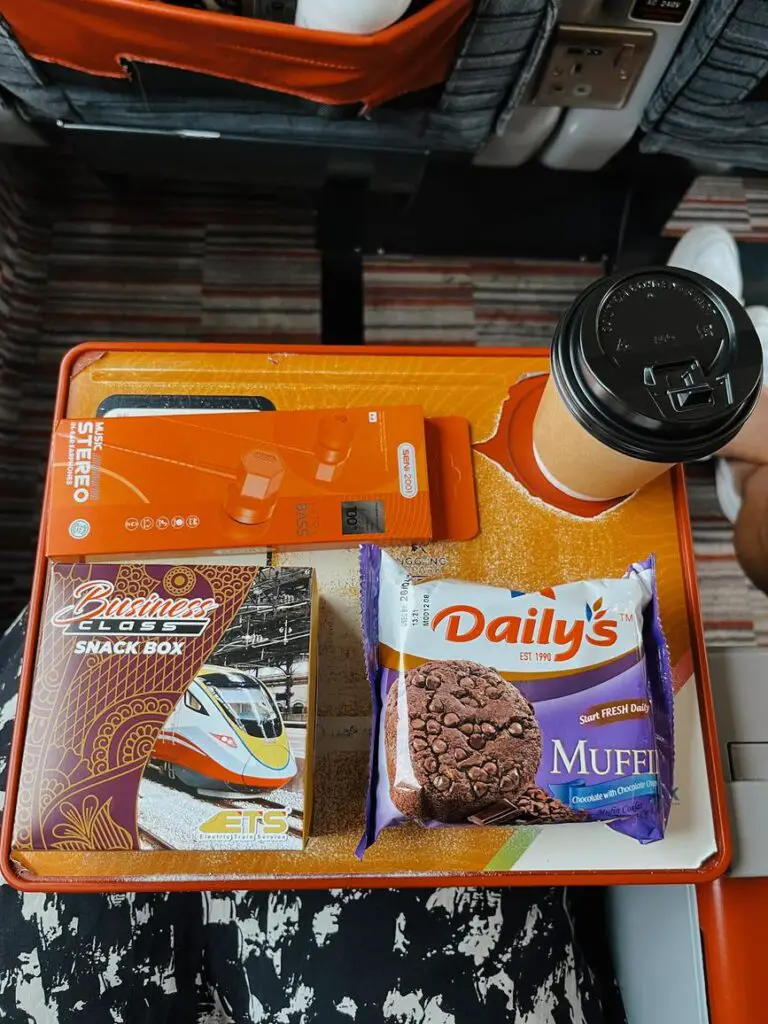 Tray table setup on a Malaysian train featuring a snack box, chocolate muffin, coffee cup, and a pair of orange earbuds, part of the business class service on one of the trains between Singapore and Bangkok.