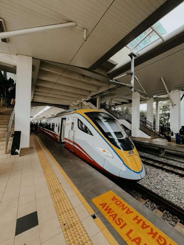 A sleek modern KTM ETS high-speed train at a platform in Malaysia, with its yellow, blue, red, and white exterior visible under a concrete station roof. A staircase and tactile paving for accessibility are seen alongside the train.