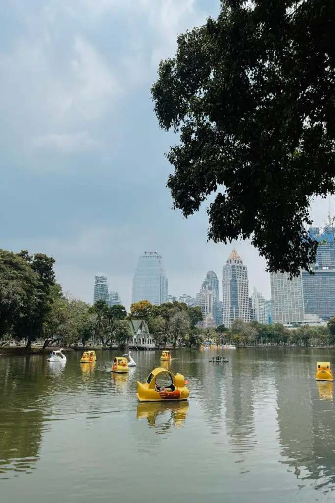 Yellow and white paddle boats shaped like ducks and swans floating on a lake in Lumphini Park, with Bangkok’s skyline in the distance.