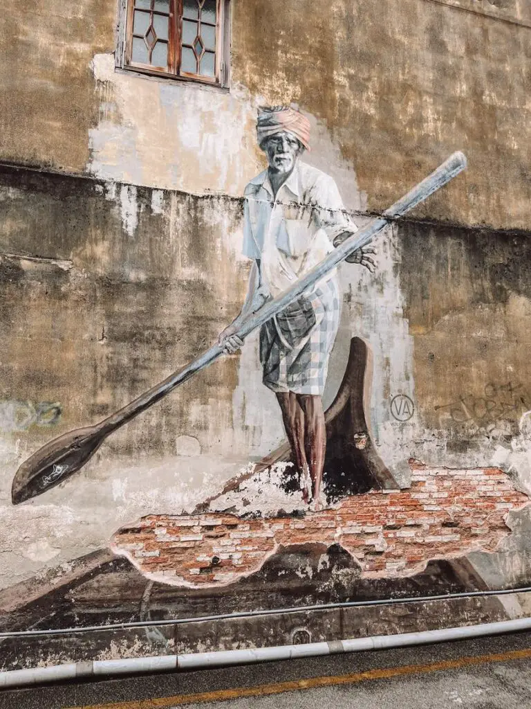 Street art mural in Penang showing an elderly man in traditional attire paddling a canoe, painted on a weathered brick and plaster wall.