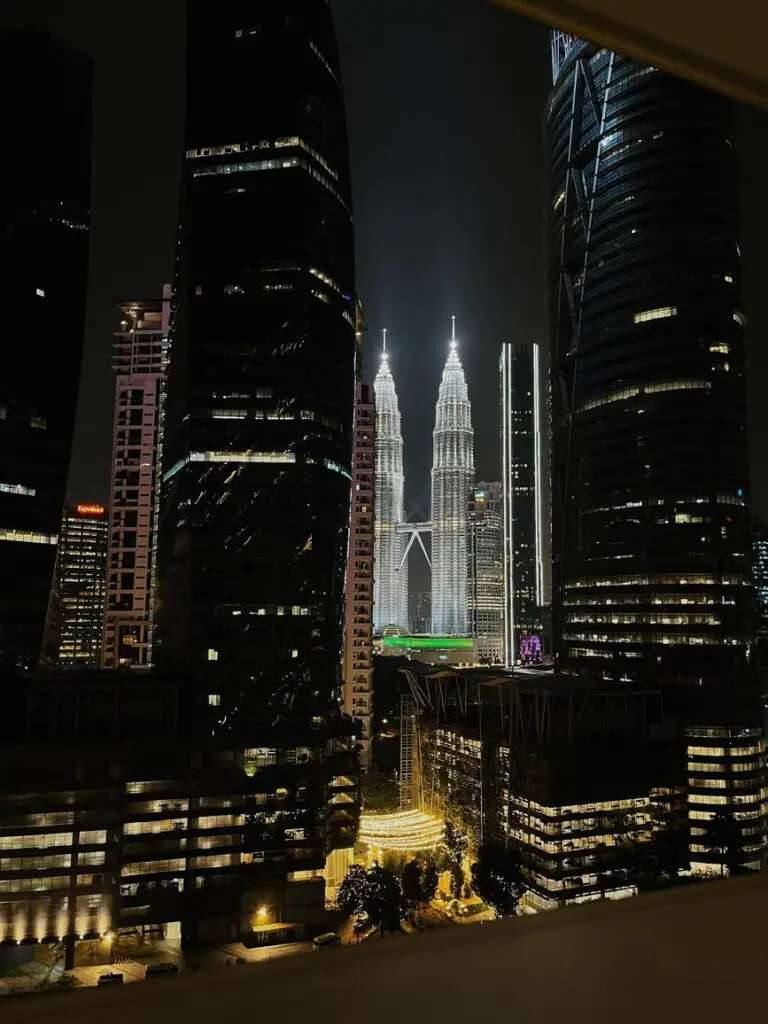 Night view of the illuminated Petronas Twin Towers framed between dark modern skyscrapers in Kuala Lumpur’s city centre.