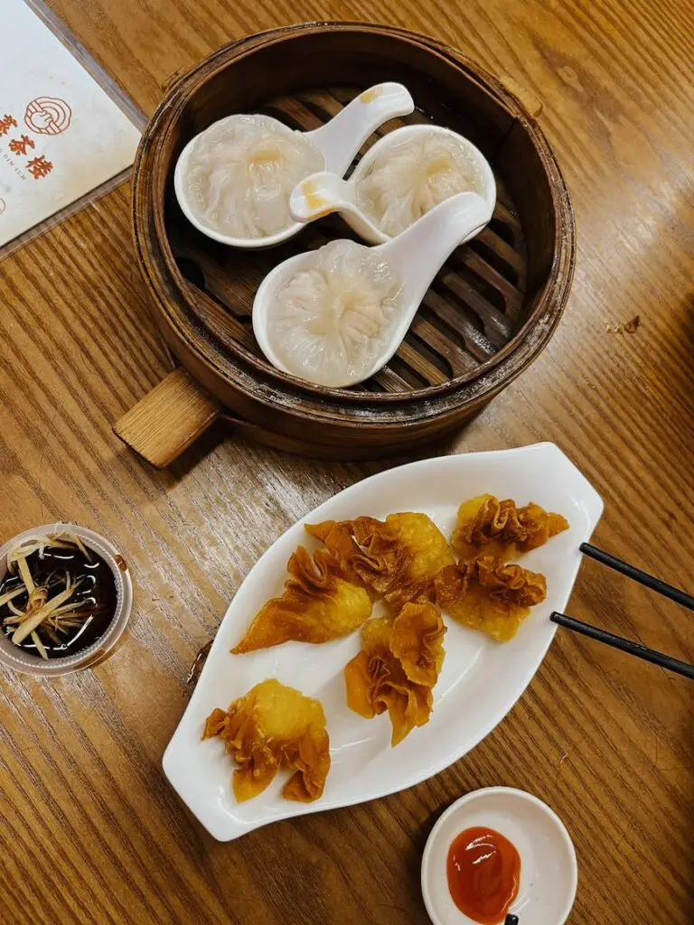 A bamboo steamer basket with three xiao long bao soup dumplings and a plate of golden fried wontons, served with dipping sauces on a wooden table.