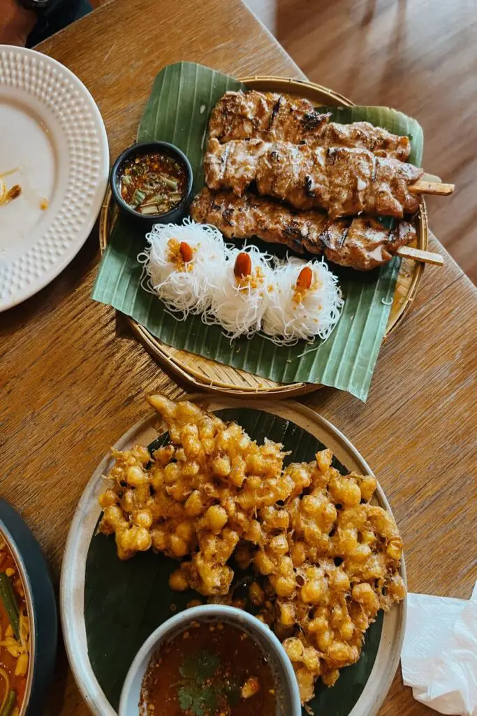 A variety of Thai dishes including grilled pork skewers with vermicelli noodles and crispy fried corn fritters, served with dipping sauces on banana leaves.