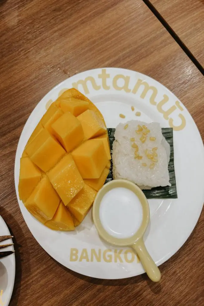 Traditional Thai dessert of ripe mango slices with sticky rice and coconut milk, served on a branded white plate with yellow text.