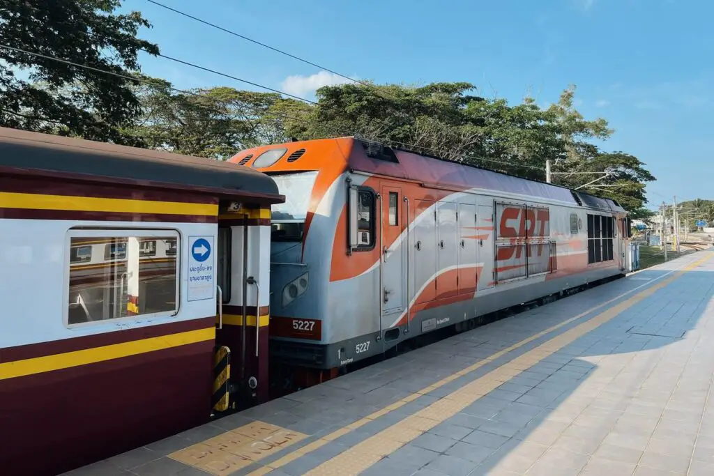 A State Railway of Thailand (SRT) locomotive at a station platform, painted red and grey with visible tracks and trees in the background.