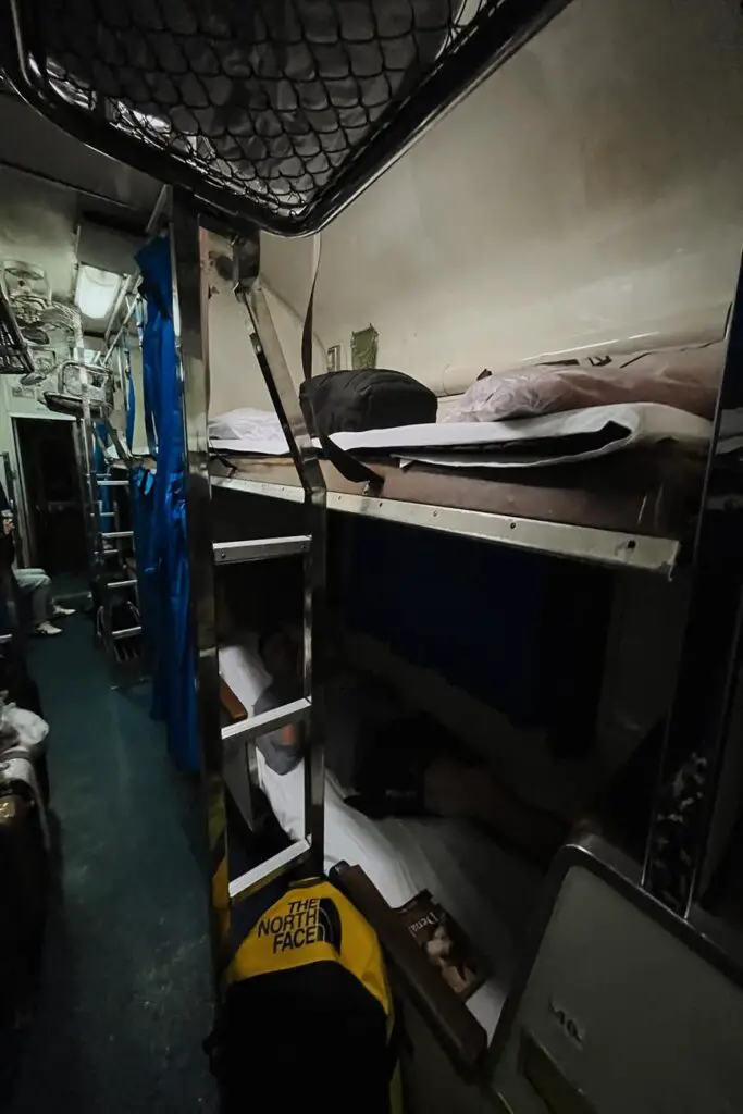 A dimly lit view of a Thai sleeper train at night showing lower and upper bunk beds with bedding, passengers resting, and a yellow backpack on the floor.