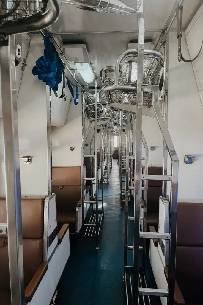 Interior view of a second-class sleeper train carriage in Thailand, showing narrow aisles, stacked metal bunk beds with storage baskets, and brown cushioned seating.