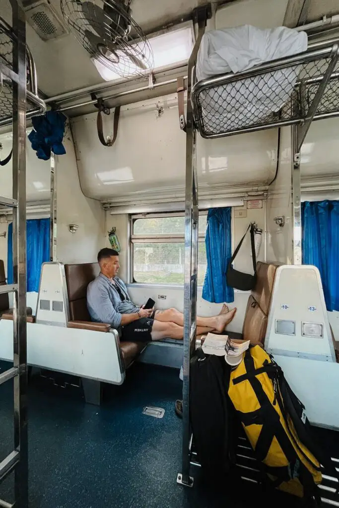 A man - the author's husband - relaxing on a brown bench seat inside a Thai sleeper train during daylight, with blue curtains, a travel backpack, and a book in view.
