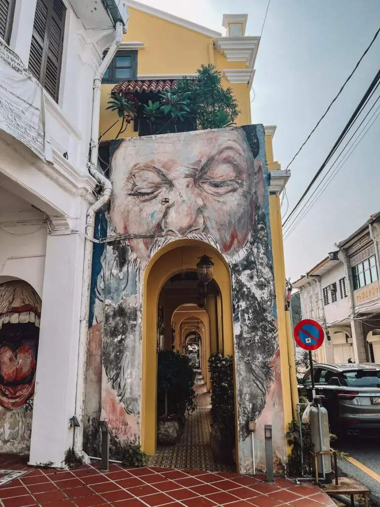 Mural of a man’s face with eyes closed, painted over a colonial-style yellow archway in Penang, Malaysia, leading to a tiled pedestrian walkway.