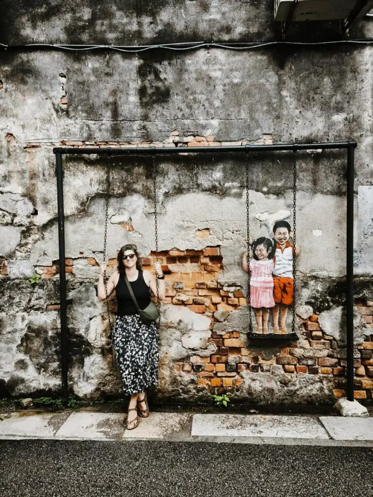 A woman - the author of this article - posing on a swing attached to a mural of two children on an adjacent swing, painted on a weathered brick and plaster wall in Penang, Malaysia. The artwork blends 3D elements with street art, creating an interactive illusion.