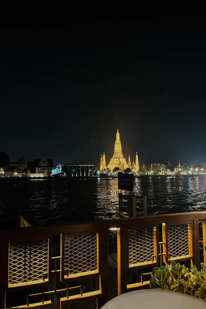 Nighttime view of the illuminated Wat Arun temple glowing gold across the Chao Phraya River, as seen from a riverside dining spot.