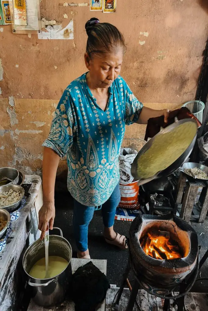 An elderly Thai woman in a blue patterned shirt cooking over a charcoal fire, preparing street food in a modest, rustic kitchen.