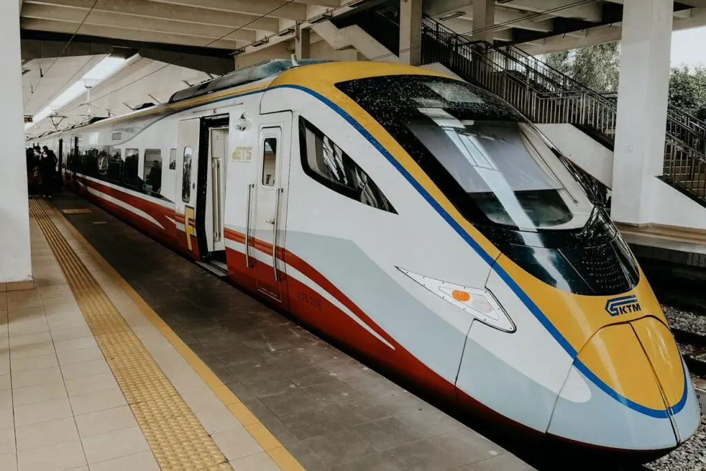 A sleek modern KTM ETS high-speed train at a platform in Malaysia, with its yellow, blue, red, and white exterior visible under a concrete station roof. A staircase and tactile paving for accessibility are seen alongside the train.