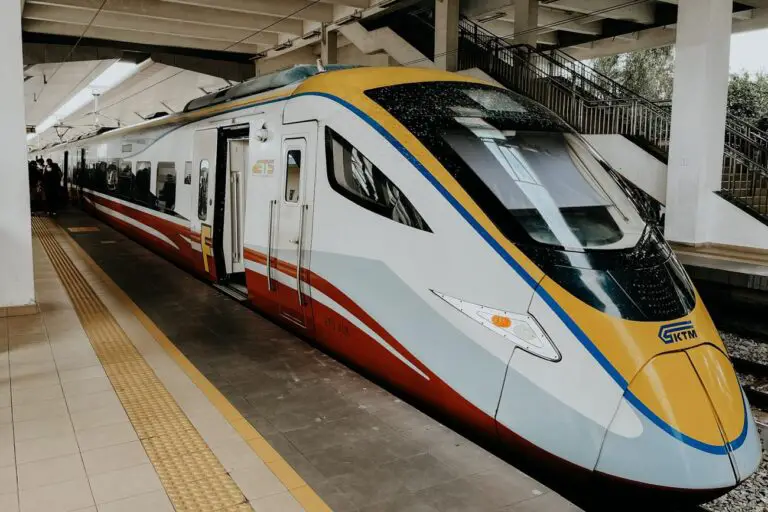 A sleek modern KTM ETS high-speed train at a platform in Malaysia, with its yellow, blue, red, and white exterior visible under a concrete station roof. A staircase and tactile paving for accessibility are seen alongside the train.