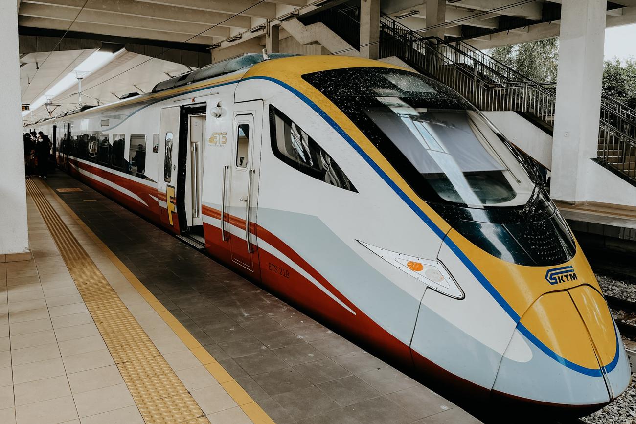 A sleek modern KTM ETS high-speed train at a platform in Malaysia, with its yellow, blue, red, and white exterior visible under a concrete station roof. A staircase and tactile paving for accessibility are seen alongside the train.