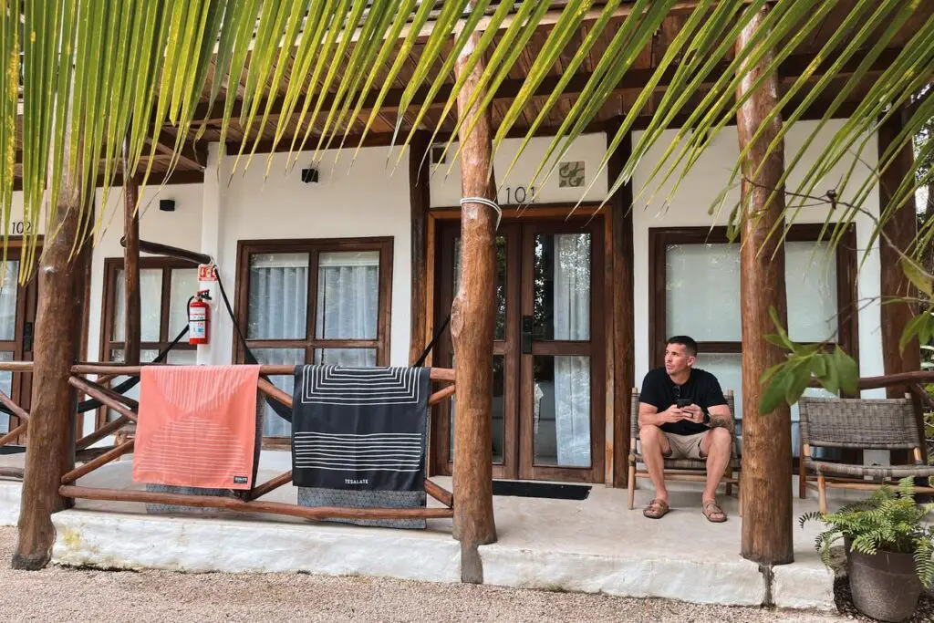 A man sits outside a simple hotel room in Bacalar, Yucatan, with beach towels hanging over a wooden railing.