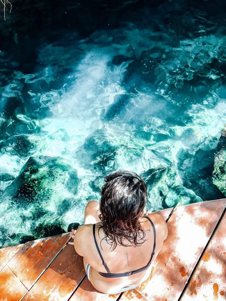 Woman - the author of this article - sitting on a wooden ledge above a clear turquoise cenote in Tulum, one of many natural swimming spots in the Yucatan.