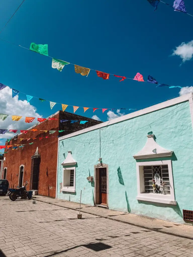 Colorful papel picado flags strung across a bright blue sky above pastel colonial buildings on Calle de los Frailes, a picturesque street in Valladolid, Mexico.