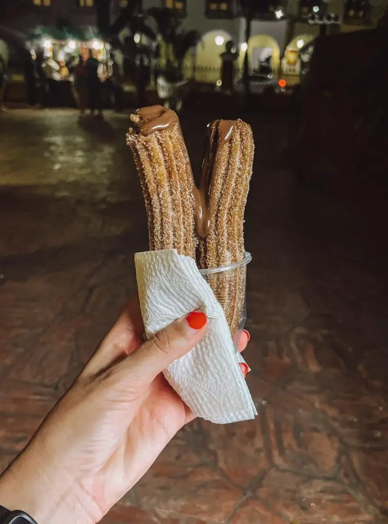 A hand holding a cup of churros drizzled with chocolate sauce in Valladolid’s main plaza at night.