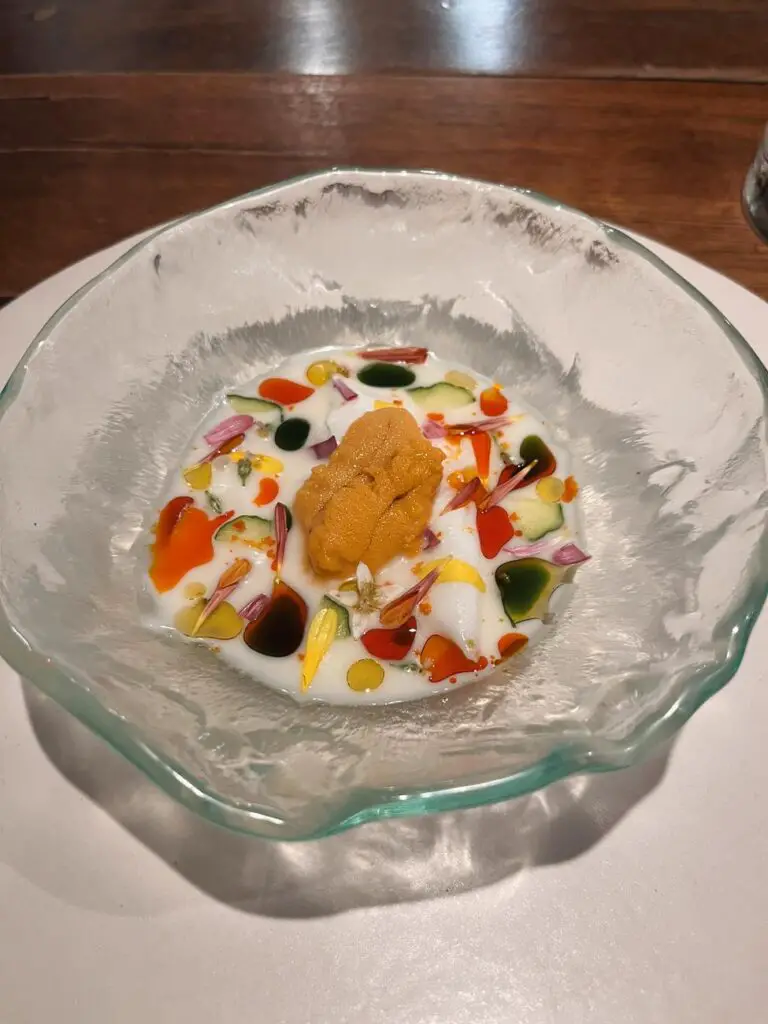 An elegant fine dining dish featuring sea urchin (uni) on a bed of creamy sauce, topped with colorful edible flowers and oil drops, served in a textured glass bowl—part of a gourmet Guadalajara food experience at Restaurant Alcalde.