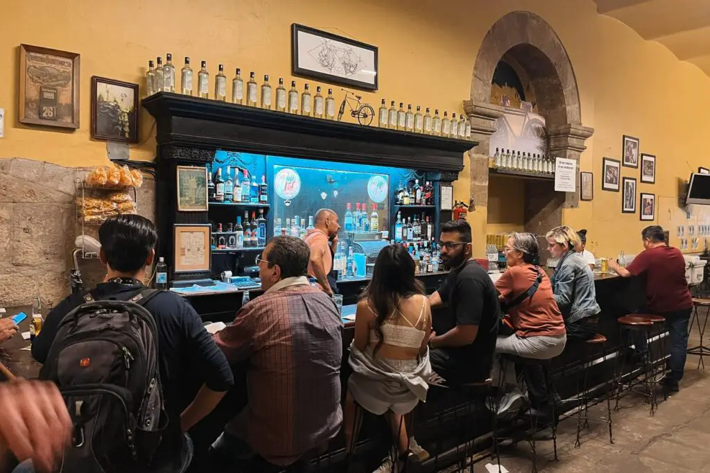 A bustling cantina in Guadalajara filled with locals and tourists enjoying drinks at the bar, known for its old-school charm and live music atmosphere.