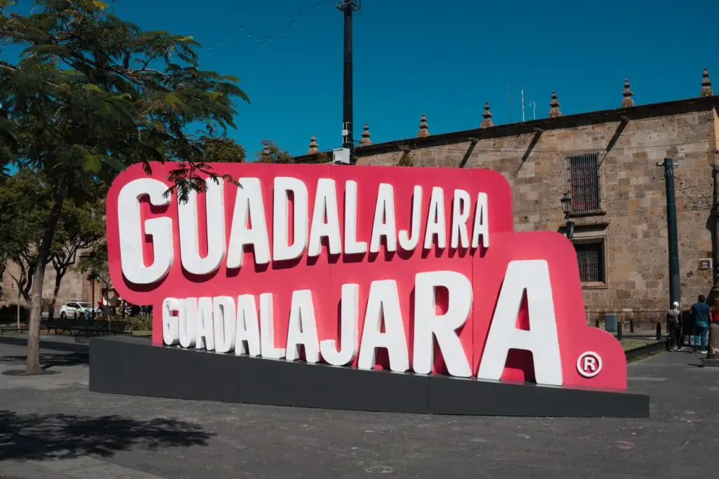 The bright red and white Guadalajara city sign in a public plaza, with colonial buildings and trees in the background—an iconic photo spot in Guadalajara.