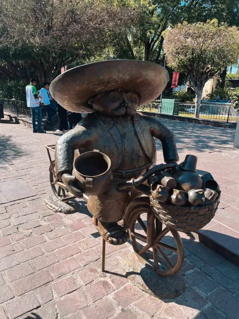 A whimsical bronze statue of a man in a sombrero riding a bicycle loaded with clay jugs, located in the charming Tlaquepaque arts district.