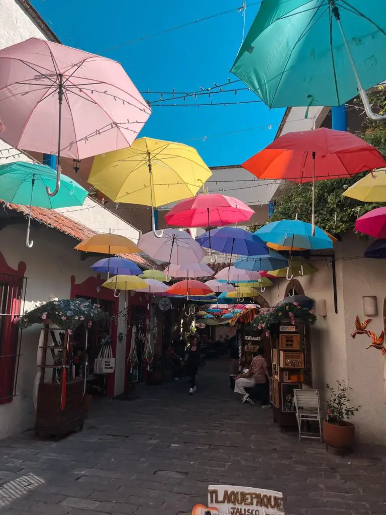 A colorful canopy of suspended umbrellas over a cobblestone street lined with artisan shops in Tlaquepaque, Jalisco—perfect for photos and shopping.